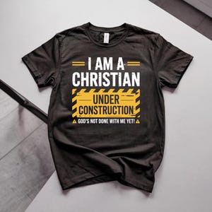 May include: Black t-shirt with the text "I AM A CHRISTIAN UNDER CONSTRUCTION" in white and yellow. The shirt also includes the phrase "GOD'S NOT DONE WITH ME YET!" in white. The design is centered on the chest.