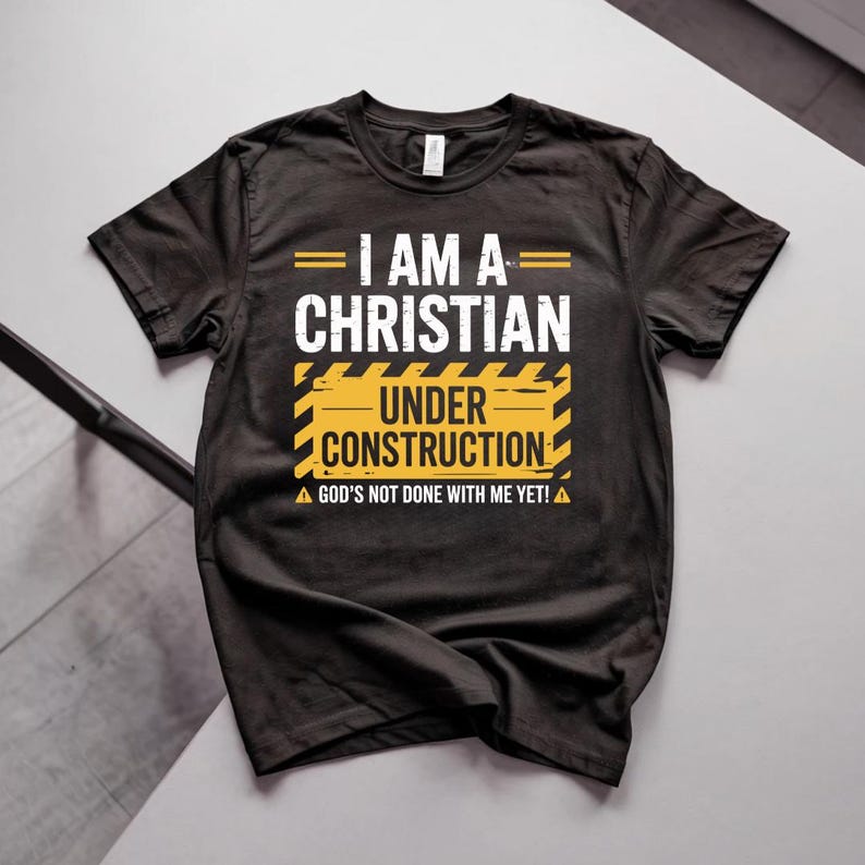 May include: Black t-shirt with the text "I AM A CHRISTIAN UNDER CONSTRUCTION" in white and yellow. The shirt also includes the phrase "GOD'S NOT DONE WITH ME YET!" in white. The design is centered on the chest.