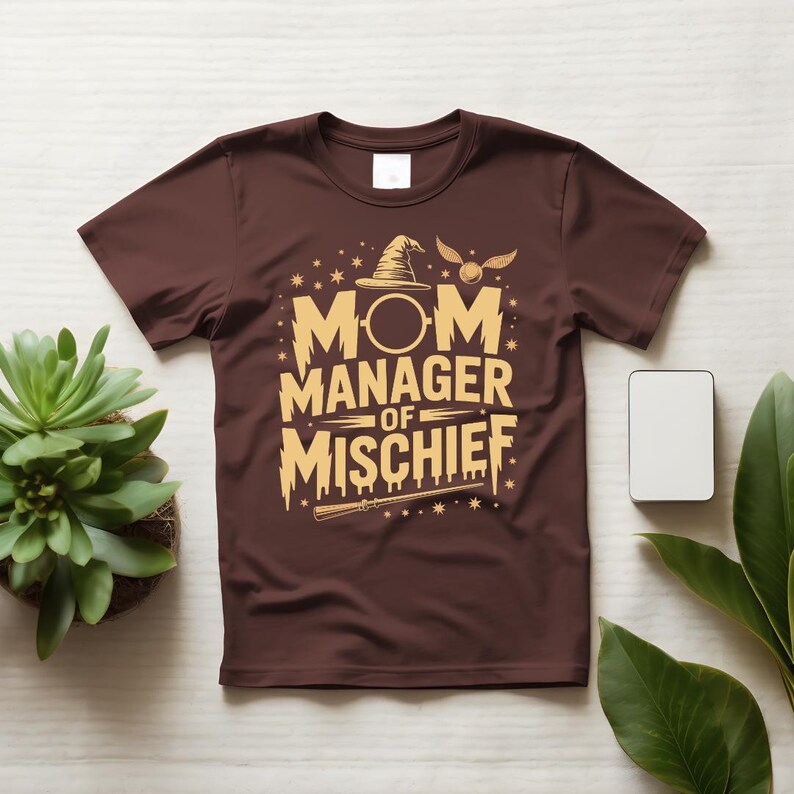 以下が含まれることがあります： 茶色のTシャツ。金色の文字で「MOM MANAGER OF MISCHIEF」と書かれています。デザインには魔法使いの帽子、星、魔法の杖が含まれています。明るい色の表面に、植物と白い長方形と一緒に表示されています。