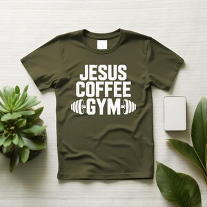 Peut inclure: T-shirt vert olive avec le texte blanc "JESUS COFFEE GYM" et un graphique d'halt&egrave;res. Le t-shirt est pr&eacute;sent&eacute; sur une surface blanche avec une petite plante succulente et un rectangle blanc.