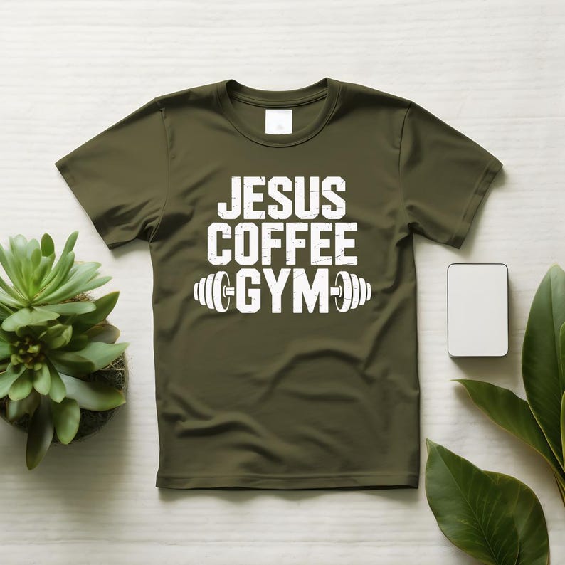 Peut inclure: T-shirt vert olive avec le texte blanc "JESUS COFFEE GYM" et un graphique d'halt&egrave;res. Le t-shirt est pr&eacute;sent&eacute; sur une surface blanche avec une petite plante succulente et un rectangle blanc.