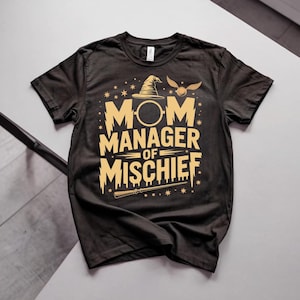 以下が含まれることがあります： 金色の文字とグラフィックが入った黒いTシャツ。「MOM MANAGER OF MISCHIEF」の文字と魔法使いの帽子、金のニワトコ、杖が描かれています。柔らかい素材で作られたシャツです。