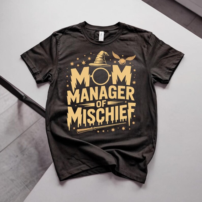 以下が含まれることがあります： 金色の文字とグラフィックが入った黒いTシャツ。「MOM MANAGER OF MISCHIEF」の文字と魔法使いの帽子、金のニワトコ、杖が描かれています。柔らかい素材で作られたシャツです。