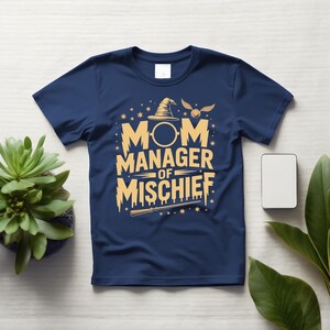 以下が含まれることがあります： 紺色のTシャツ。金色の文字で「MOM MANAGER OF MISCHIEF」と書かれています。デザインには魔法使いの帽子、金のスニッチ、魔法の杖が含まれています。クルーネック、半袖です。