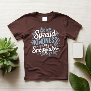 May include: A brown t-shirt featuring the text "Spread KINDNESS Like Snowflakes" in white and light blue, with snowflake designs. The shirt is shown on a light wooden surface.