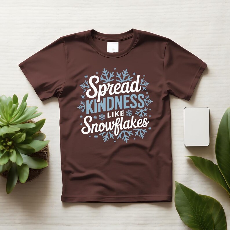 May include: A brown t-shirt featuring the text "Spread KINDNESS Like Snowflakes" in white and light blue, with snowflake designs. The shirt is shown on a light wooden surface.