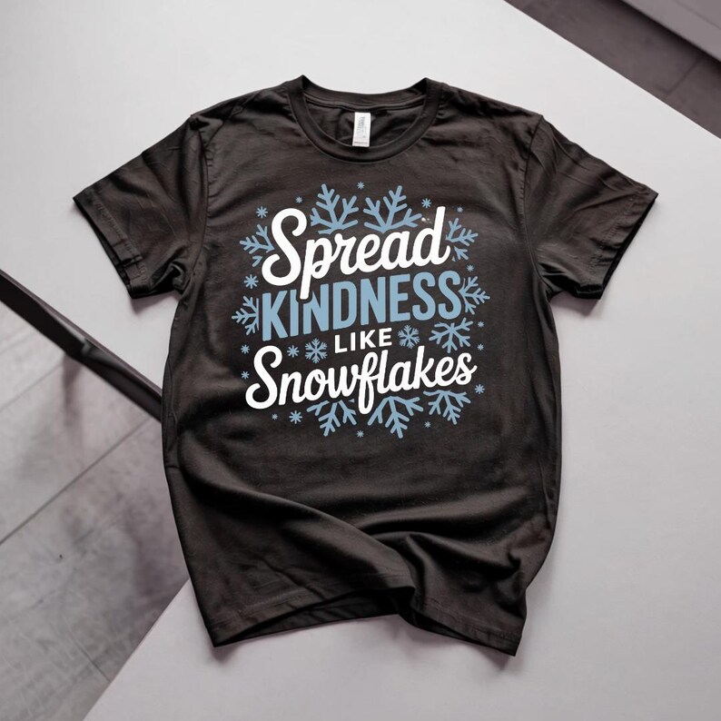 May include: A dark grey t-shirt with the text "Spread KINDNESS like Snowflakes" in white and light blue. The design features snowflakes around the text. The shirt is a crew neck style.