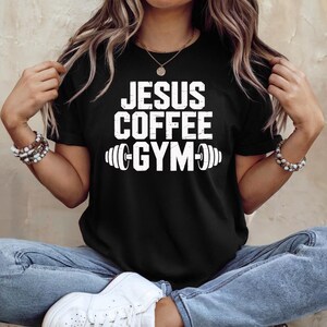 Peut inclure: T-shirt noir avec l'inscription blanche "JESUS COFFEE GYM" et un graphique de barre d'halt&egrave;res. Le t-shirt est un col rond et est fait d'une mati&egrave;re douce. Le t-shirt est port&eacute; par une personne portant un jean bleu.