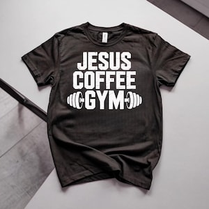 Peut inclure: T-shirt noir avec l'inscription blanche "JESUS COFFEE GYM" et un motif d'halt&egrave;re. Le t-shirt est de style col rond et semble &ecirc;tre fait d'une mati&egrave;re douce. Le texte est centr&eacute; sur le devant du t-shirt.