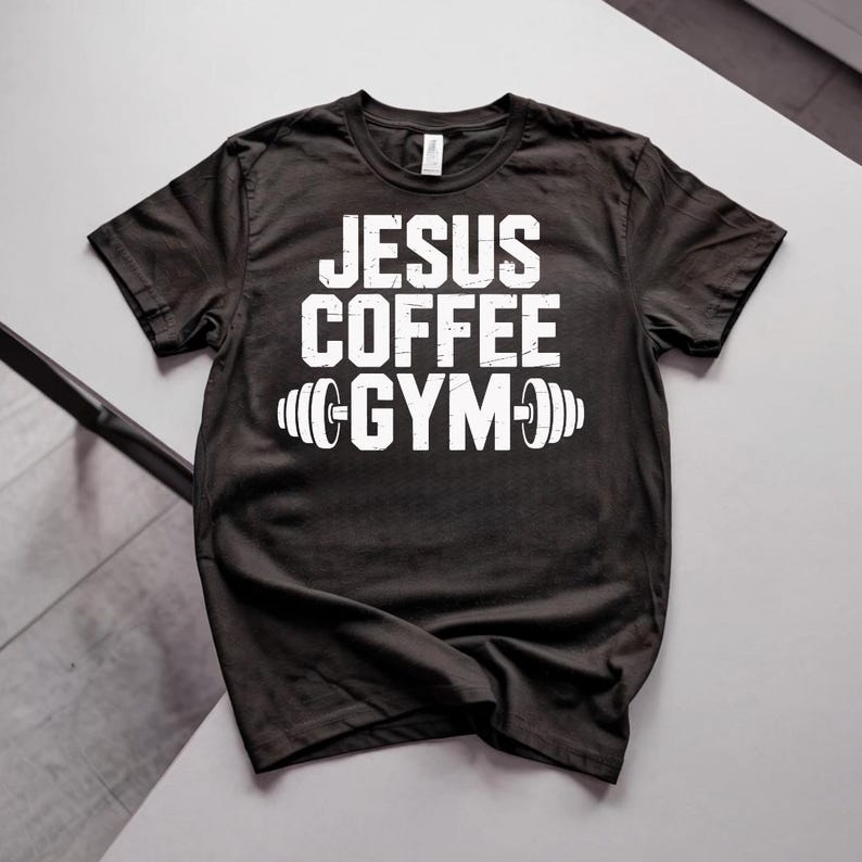 Peut inclure: T-shirt noir avec l'inscription blanche "JESUS COFFEE GYM" et un motif d'halt&egrave;re. Le t-shirt est de style col rond et semble &ecirc;tre fait d'une mati&egrave;re douce. Le texte est centr&eacute; sur le devant du t-shirt.