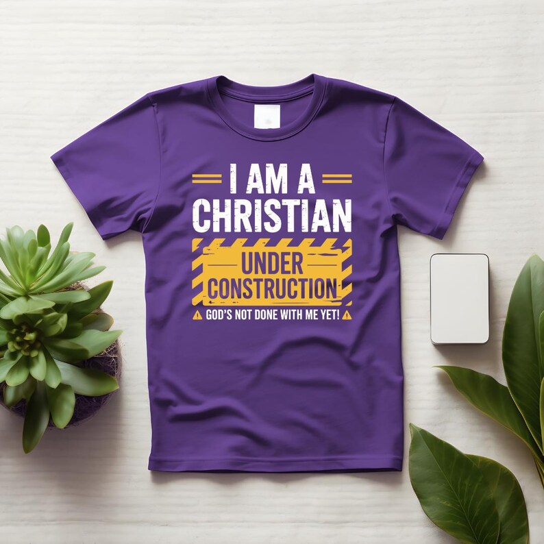 May include: A purple t-shirt with the text "I AM A CHRISTIAN UNDER CONSTRUCTION" in white and yellow. The shirt also includes the text "GOD'S NOT DONE WITH ME YET!" in white. The shirt is displayed on a white surface.