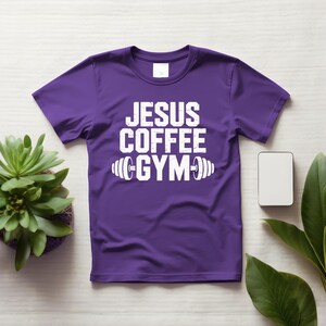 Peut inclure: T-shirt violet avec l'inscription blanche "JESUS COFFEE GYM" et un halt&egrave;re. Le t-shirt a des manches courtes et un col rond. Une petite plante succulente et un rectangle blanc vierge sont visibles.