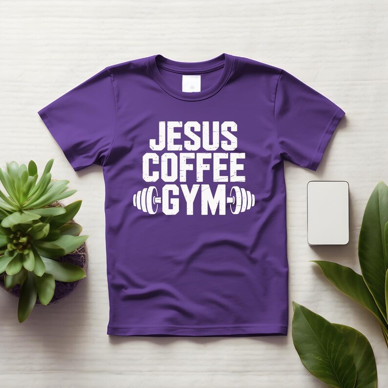 Peut inclure: T-shirt violet avec l'inscription blanche "JESUS COFFEE GYM" et un halt&egrave;re. Le t-shirt a des manches courtes et un col rond. Une petite plante succulente et un rectangle blanc vierge sont visibles.
