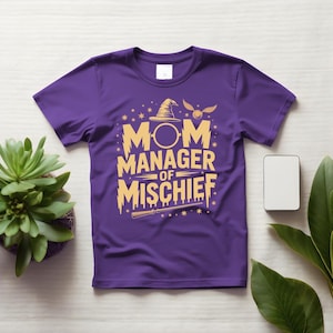 以下が含まれることがあります： 紫色のTシャツ。金色の文字で「MOM MANAGER OF MISCHIEF」と書かれています。デザインには魔法使いの帽子、金のスニッチ、魔法の杖が含まれています。Tシャツは、植物と電話が置かれた白い表面に表示されています。