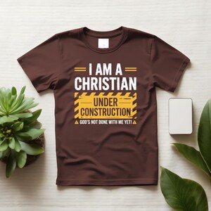 May include: A brown t-shirt with the text "I AM A CHRISTIAN UNDER CONSTRUCTION" in white and yellow. The shirt also includes the phrase "GOD'S NOT DONE WITH ME YET!" in white. The shirt is displayed on a white surface with plants.