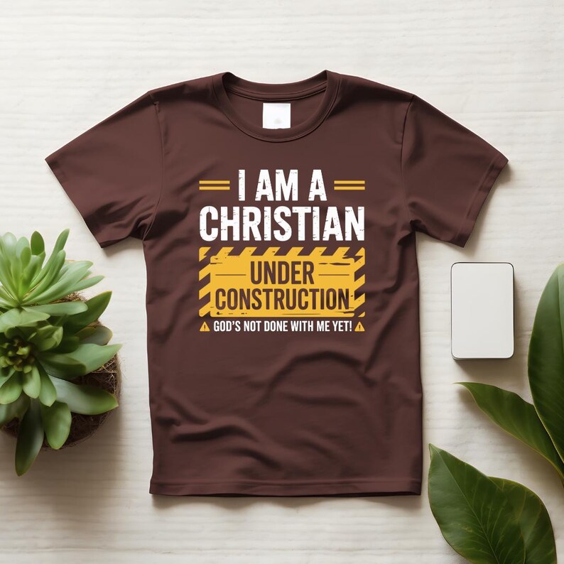 May include: A brown t-shirt with the text "I AM A CHRISTIAN UNDER CONSTRUCTION" in white and yellow. The shirt also includes the phrase "GOD'S NOT DONE WITH ME YET!" in white. The shirt is displayed on a white surface with plants.