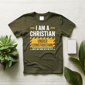 May include: Olive green t-shirt with the text "I AM A CHRISTIAN UNDER CONSTRUCTION" in white and yellow. The shirt also includes the phrase "GOD'S NOT DONE WITH ME YET!" in white. The design is centered on the front of the shirt.