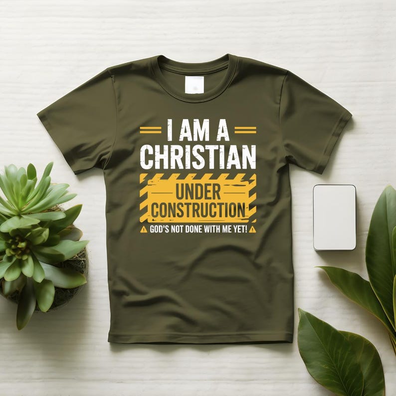 May include: Olive green t-shirt with the text "I AM A CHRISTIAN UNDER CONSTRUCTION" in white and yellow. The shirt also includes the phrase "GOD'S NOT DONE WITH ME YET!" in white. The design is centered on the front of the shirt.
