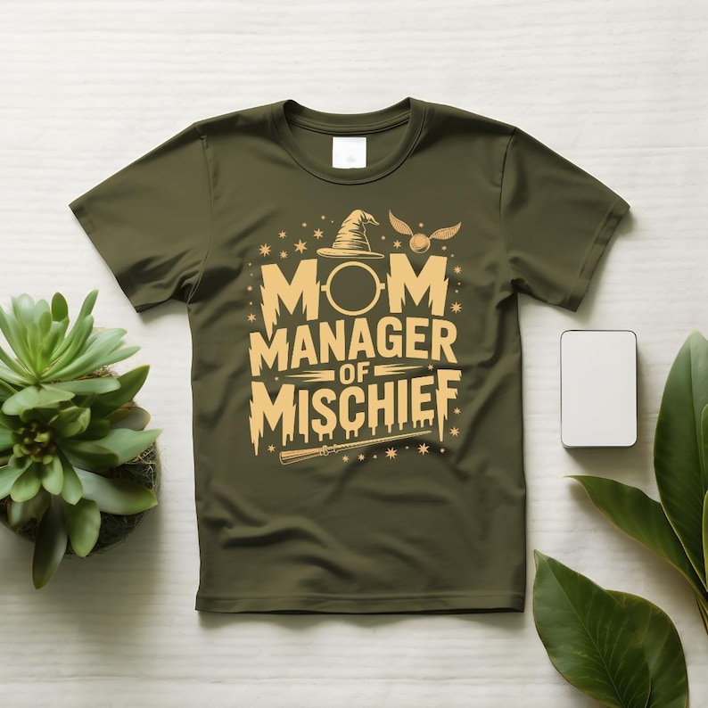 以下が含まれることがあります： オリーブグリーンのTシャツ。金色の文字で「MOM MANAGER OF MISCHIEF」と書かれています。デザインには魔法使いの帽子、杖、星が含まれています。Tシャツは、植物と白い長方形のある白い表面に表示されています。