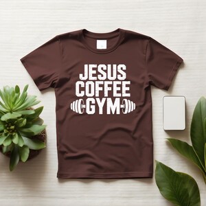 Peut inclure: Un t-shirt marron avec l'inscription blanche "JESUS COFFEE GYM" et un motif d'halt&egrave;re. Le t-shirt est pr&eacute;sent&eacute; sur une surface claire, avec une petite plante et un rectangle blanc vierge en arri&egrave;re-plan.