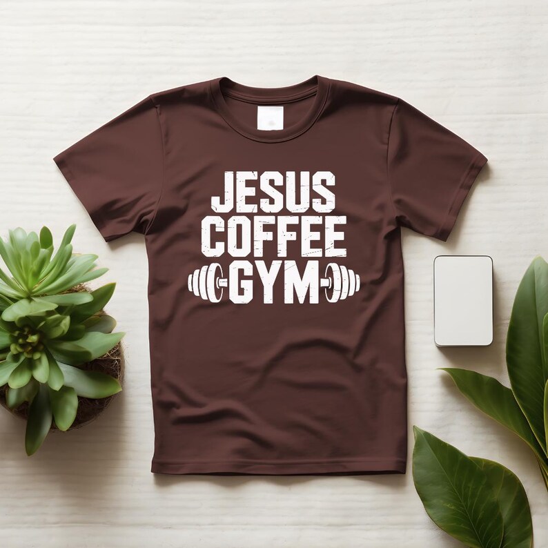 Peut inclure: Un t-shirt marron avec l'inscription blanche "JESUS COFFEE GYM" et un motif d'halt&egrave;re. Le t-shirt est pr&eacute;sent&eacute; sur une surface claire, avec une petite plante et un rectangle blanc vierge en arri&egrave;re-plan.