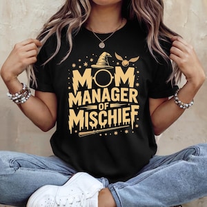 以下が含まれることがあります： 黒いTシャツ。金色の文字で「MOM MANAGER OF MISCHIEF」と書かれています。デザインには魔法使いの帽子、金のスニッチ、杖が含まれています。Tシャツはクルーネックで半袖です。