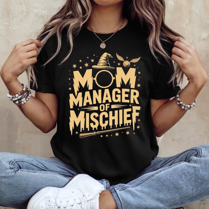 以下が含まれることがあります： 黒いTシャツ。金色の文字で「MOM MANAGER OF MISCHIEF」と書かれています。デザインには魔法使いの帽子、金のスニッチ、杖が含まれています。Tシャツはクルーネックで半袖です。
