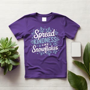 May include: A purple t-shirt with the slogan "Spread Kindness Like Snowflakes" in white and light blue, surrounded by snowflake designs. The shirt is shown on a light surface, with a small plant and a white rectangle nearby.