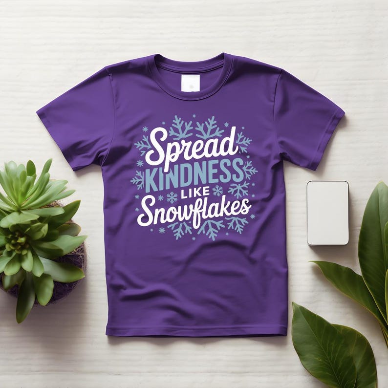 May include: A purple t-shirt with the slogan "Spread Kindness Like Snowflakes" in white and light blue, surrounded by snowflake designs. The shirt is shown on a light surface, with a small plant and a white rectangle nearby.