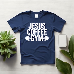 Peut inclure: T-shirt bleu marine avec l'inscription blanche "JESUS COFFEE GYM" et un halt&egrave;re. Le t-shirt est pos&eacute; sur une surface en bois blanc, avec une petite plante et un rectangle blanc.