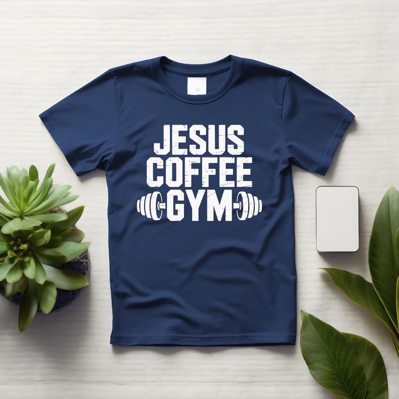 Peut inclure: T-shirt bleu marine avec l'inscription blanche "JESUS COFFEE GYM" et un halt&egrave;re. Le t-shirt est pos&eacute; sur une surface en bois blanc, avec une petite plante et un rectangle blanc.