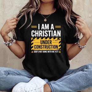 May include: Black t-shirt with the text "I AM A CHRISTIAN UNDER CONSTRUCTION". The design includes yellow and black construction-themed elements and the phrase "GOD'S NOT DONE WITH ME YET!"