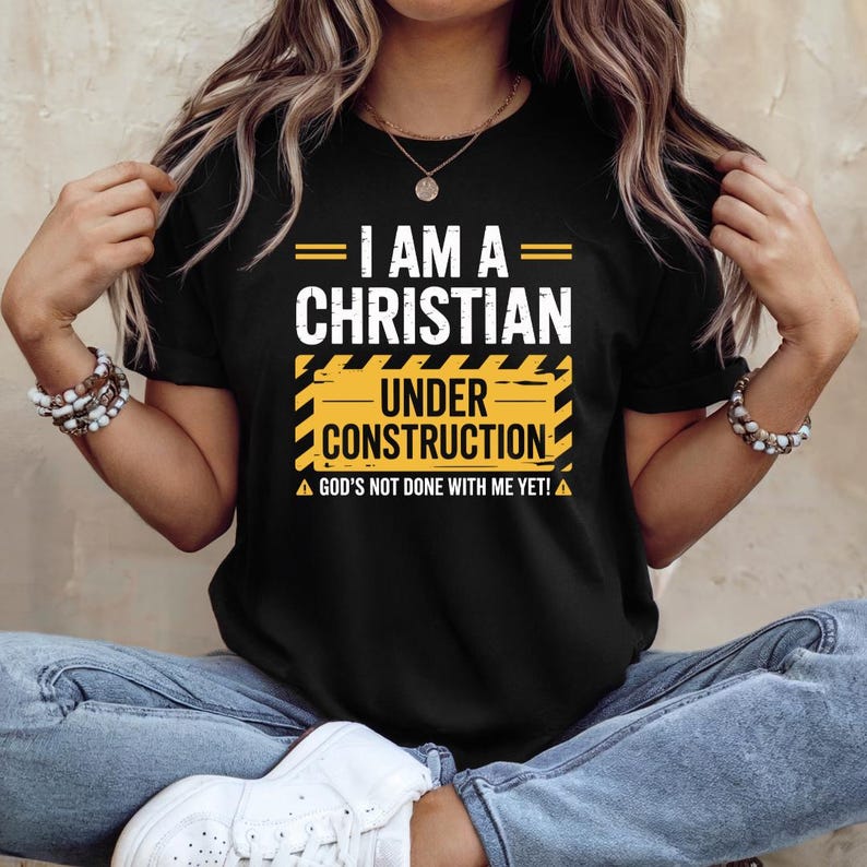 May include: Black t-shirt with the text "I AM A CHRISTIAN UNDER CONSTRUCTION". The design includes yellow and black construction-themed elements and the phrase "GOD'S NOT DONE WITH ME YET!"