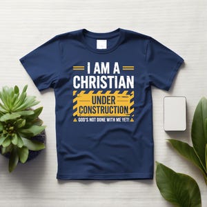 May include: Navy blue t-shirt with the text "I AM A CHRISTIAN UNDER CONSTRUCTION." The text is in white and yellow, with a construction-themed design. The shirt also includes the phrase "GOD'S NOT DONE WITH ME YET!"