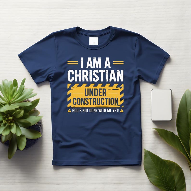 May include: Navy blue t-shirt with the text "I AM A CHRISTIAN UNDER CONSTRUCTION." The text is in white and yellow, with a construction-themed design. The shirt also includes the phrase "GOD'S NOT DONE WITH ME YET!"