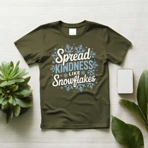 May include: An olive green t-shirt featuring the text "Spread KINDNESS like Snowflakes" in white and light blue, with snowflake designs. The shirt is shown on a light surface, accompanied by a small plant and a white rectangle.