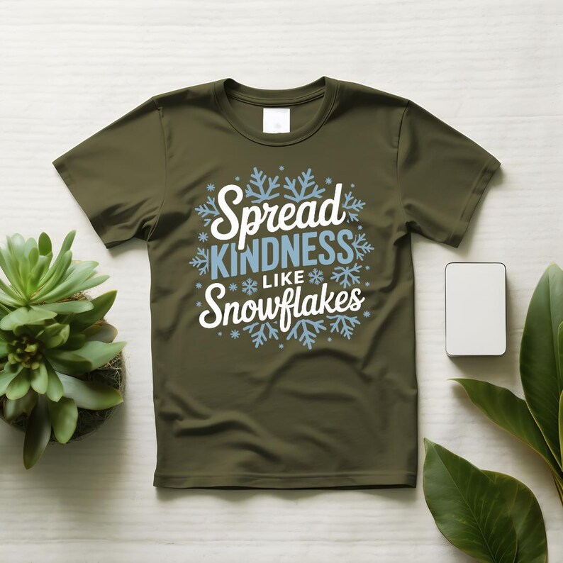 May include: An olive green t-shirt featuring the text "Spread KINDNESS like Snowflakes" in white and light blue, with snowflake designs. The shirt is shown on a light surface, accompanied by a small plant and a white rectangle.