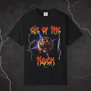 May include: Black t-shirt featuring a graphic print of a tiger with glowing eyes and lightning bolts. The text "Eye of the Tiger" is printed in a metal font above the tiger and the text "Tiger" is printed in a metal font below the tiger.