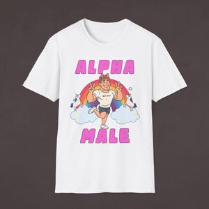 Pode incluir: Camiseta branca com as palavras "ALPHA MALE" em letras rosa. A camisa apresenta uma ilustração de desenho animado de um homem com um arco-íris, unicórnios e nuvens.