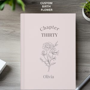 May include: A pale pink journal featuring the text "Chapter THIRTY" and the name "Olivia" beneath a floral design. The words "CUSTOM BIRTH FLOWER" are at the top. The journal rests on a wooden surface.