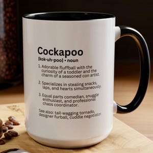 May include: White ceramic mug with a black interior and handle. The mug features the definition of "Cockapoo" with humorous descriptions. The text includes the word "Cockapoo" and its pronunciation, along with three numbered characteristics.