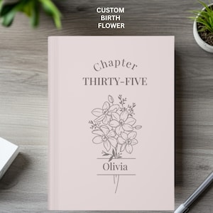 May include: A light pink journal with the text "Chapter THIRTY-FIVE" and "Olivia" below a floral illustration. The top of the journal reads "CUSTOM BIRTH FLOWER".