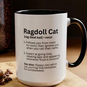Peut inclure: Un mug en céramique blanc avec un intérieur et une anse noirs, présentant un texte imprimé en noir qui définit le Ragdoll Cat comme un nom, incluant des descriptions humoristiques de son comportement et une liste de termes associés.