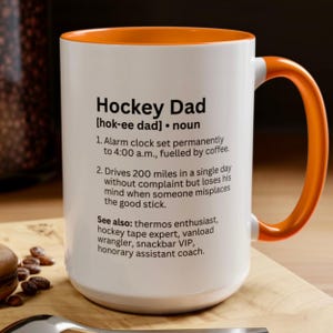 K&ouml;nnte beinhalten: Wei&szlig;er Keramikbecher mit orangefarbenem Innenraum und Griff. Der Becher zeigt den Text "Hockey Dad" mit einer Definition und humorvollen Beschreibungen. Ein perfektes Geschenk f&uuml;r Hockey-Enthusiasten.