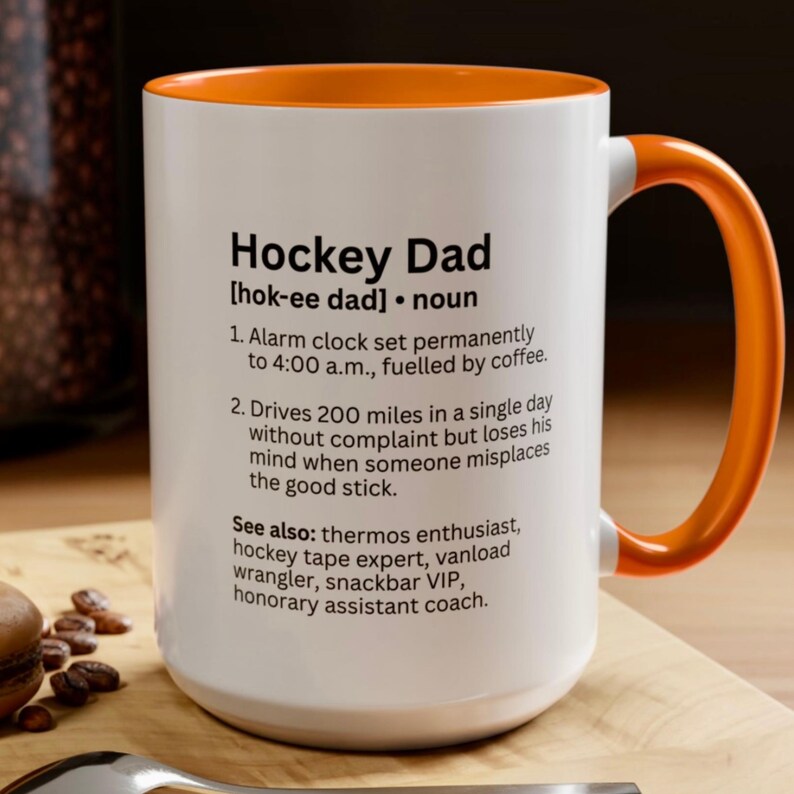 K&ouml;nnte beinhalten: Wei&szlig;er Keramikbecher mit orangefarbenem Innenraum und Griff. Der Becher zeigt den Text "Hockey Dad" mit einer Definition und humorvollen Beschreibungen. Ein perfektes Geschenk f&uuml;r Hockey-Enthusiasten.