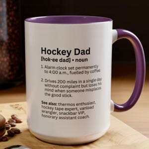 K&ouml;nnte beinhalten: Wei&szlig;er Keramikbecher mit lila Innenraum und Henkel. Der Becher zeigt den Text "Hockey Dad" mit einer Definition, einschlie&szlig;lich der S&auml;tze "Wecker" und "322 Kilometer". Der Becher steht auf einer Holzoberfl&auml;che.
