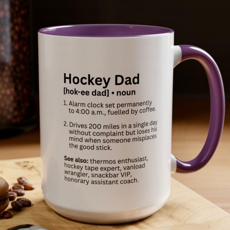 K&ouml;nnte beinhalten: Wei&szlig;er Keramikbecher mit lila Innenraum und Henkel. Der Becher zeigt den Text "Hockey Dad" mit einer Definition, einschlie&szlig;lich der S&auml;tze "Wecker" und "322 Kilometer". Der Becher steht auf einer Holzoberfl&auml;che.