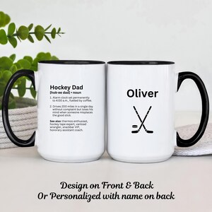 K&ouml;nnte beinhalten: Wei&szlig;er Keramikbecher mit schwarzem Griff und Rand. Eine Seite zeigt die Definition von "Hockey Dad" und die andere Seite den Namen "Oliver" mit Hockeyschl&auml;gern und einem Puck. Der Becher ist ein Geschenk f&uuml;r einen Hockey-Enthusiasten.