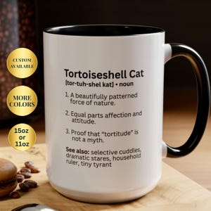 May include: A white ceramic mug with a black interior and handle, featuring black text that defines a Tortoiseshell Cat as a patterned force of nature, noting that tortitude is not a myth, and listing see also terms like tiny tyrant.