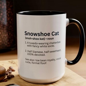 May include: A white ceramic mug with a black interior and handle. The front features black text defining a Snowshoe Cat as a tuxedo-wearing chatterbox with white socks, half Siamese, half sweetheart, 100% devoted, and also toe bean royalty.